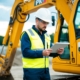 An excavator operator wearing a hard hat and reflective vest performs a pre-operation safety check on the equipment at a construction site